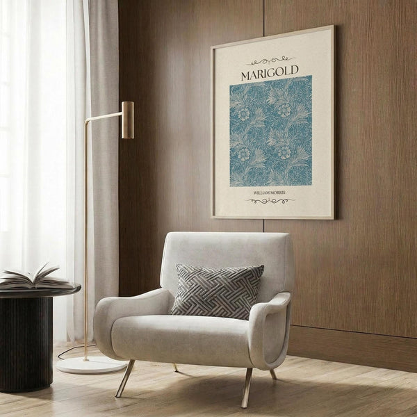 May include: Framed poster with blue floral pattern and "MARIGOLD" and "WILLIAM MORRIS" text displayed on wood-paneled wall above beige upholstered chair with geometric patterned pillow. Modern brass floor lamp with white base stands nearby next to dark round side table with open book. Neutral toned room setting with natural lighting from window.