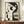 May include: Abstract geometric artwork featuring a stylized woman's profile in black and beige tones, displayed in a thin wooden frame on a walnut credenza. The minimalist composition shows curved and angular shapes forming facial features, with circular and linear elements. A small white ceramic vase and stacked books sit nearby on the mid-century modern furniture against cream wainscoted walls.
