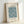 May include: Framed botanical art print featuring William Morris's Marigold pattern in blue-green tones with decorative flowers and flowing foliage design, displayed in light wood frame against white wall corner, casting soft shadows