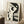 May include: Abstract minimalist poster featuring a stylized woman's profile in black and cream tones, displayed in a light wood frame leaning against white paneled walls. The artwork shows geometric shapes forming a feminine silhouette with closed eyes and flowing hair. Scene includes a coffee cup on a wooden block side table and textured beige throw blanket, creating a cozy modern living space with natural lighting.