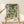 May include: Hands holding a framed botanical art print featuring lush floral patterns with cream peonies, green foliage, and orange accent flowers on a dark background. The wooden-framed poster displays "WILLIAM MORRIS" text at bottom and rests on a light wood shelf next to a small wrapped gift with twine bow against a cream wall.