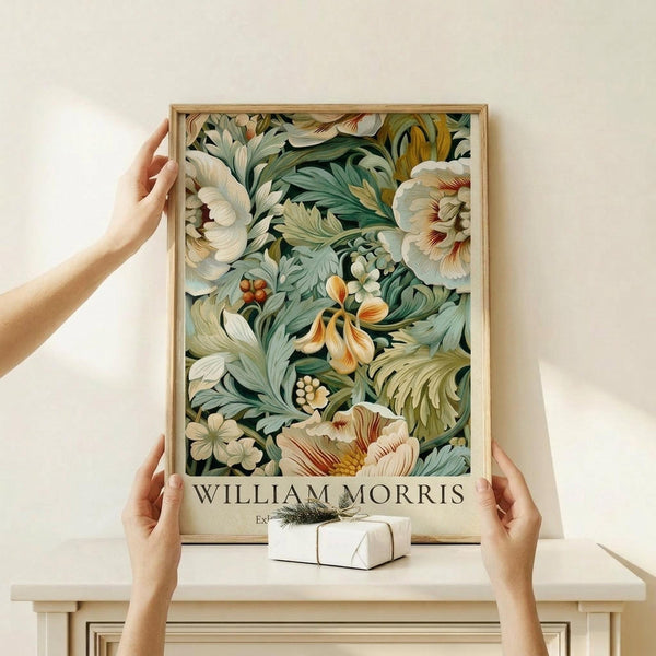 May include: Hands holding a framed botanical art print featuring lush floral patterns with cream peonies, green foliage, and orange accent flowers on a dark background. The wooden-framed poster displays "WILLIAM MORRIS" text at bottom and rests on a light wood shelf next to a small wrapped gift with twine bow against a cream wall.