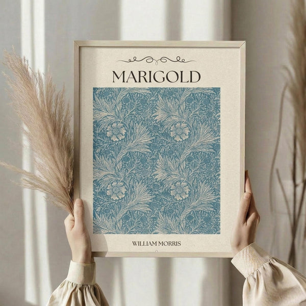 May include: Hands holding a framed botanical art print featuring William Morris's marigold pattern in blue-green tones with intricate floral and leaf designs. Light wood frame against cream background with dried pampas grass visible on left side. Person wearing beige ruffled sleeve top in soft natural lighting near window.