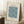 May include: Framed poster with cream background featuring blue William Morris floral pattern with marigold motifs and decorative text, displayed in light wood frame leaning against white wall corner, with coffee cup on wooden block and beige blanket nearby on white textured rug