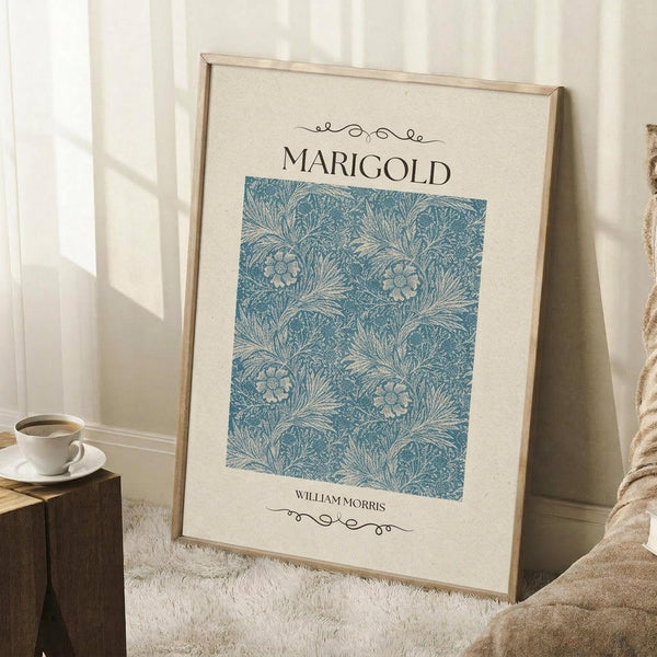 May include: Framed poster with cream background featuring blue William Morris floral pattern with marigold motifs and decorative text, displayed in light wood frame leaning against white wall corner, with coffee cup on wooden block and beige blanket nearby on white textured rug
