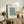 May include: Framed botanical art print with blue floral pattern displayed on rustic wooden sideboard alongside fern plant, books, and ceramic vase in warm neutral living room setting with beige upholstered chair
