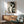 May include: Abstract geometric poster featuring a stylized woman's profile in black and cream tones, displayed in thin wood frame above mid-century modern walnut credenza with white ceramic lamp. Sage green upholstered armchair with wooden arms sits nearby on hardwood floor. Natural sunlight creates dramatic shadows on white wall in minimalist living room setting.