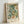 May include: Framed botanical art print featuring William Morris-inspired floral design with cream peonies, sage green foliage, and orange accent flowers against dark background. Light wood frame with white matting displays exhibition-style text at bottom. Poster leans against white wall on light surface, showing natural lighting and shadows.