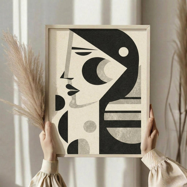 May include: Person in beige clothing holding framed abstract art print featuring stylized woman's profile in black and cream geometric shapes. Light wood frame, neutral wall background, dried pampas grass held in other hand, soft natural lighting creates minimalist home decor scene.