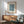 May include: Framed William Morris marigold pattern print in blue-green tones hanging above a wooden sideboard with white ceramic lamp. Mid-century modern sage green armchair with wooden arms sits beside a round coffee table. Natural sunlight creates shadows across white walls in this minimalist living room setting.