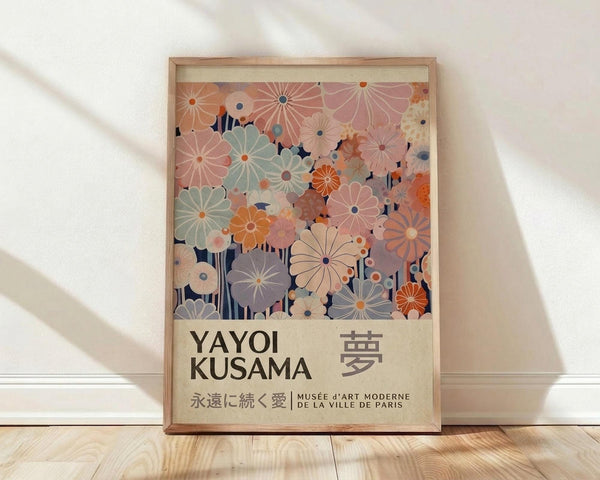 May include: Framed art print featuring colorful floral pattern with overlapping daisy-like flowers in soft pastels - mint green, coral pink, orange, and purple blooms against navy background. Text reads "YAYOI KUSAMA" with Japanese characters and "永遠に続く愛 MUSÉE D'ART MODERNE DE LA VILLE DE PARIS" on cream background. Natural wood frame leaning against white wall on light wooden floor with soft window light shadows.