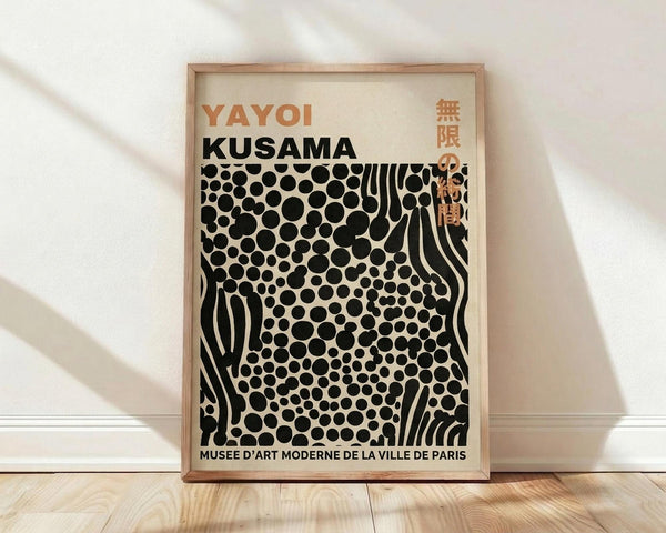 May include: A framed art print leaning against a white wall on light wood flooring. The poster features black polka dots and flowing wavy lines on a cream background, with "YAYOI KUSAMA" in orange and black text at the top and "MUSEE D'ART MODERNE DE LA VILLE DE PARIS" at the bottom. The thin wooden frame casts soft shadows on the wall from natural lighting.