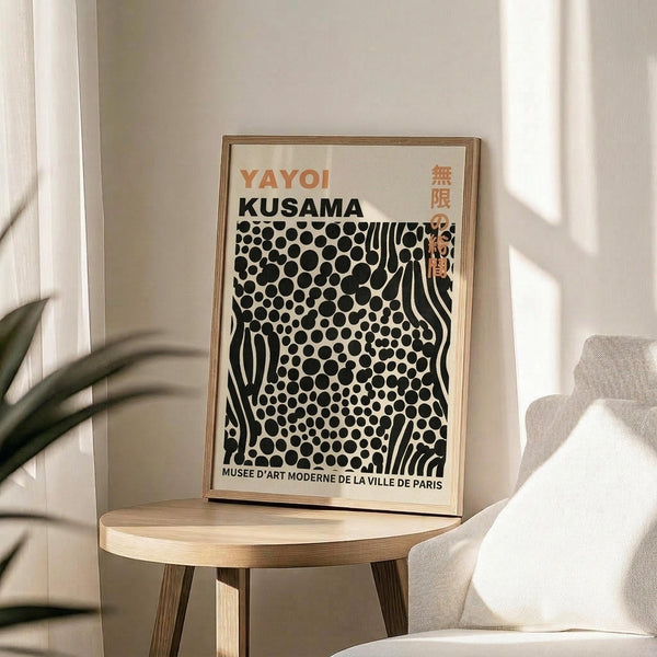 May include: Framed poster featuring Yayoi Kusama's black polka dot pattern with wavy lines on cream background, displayed on round wooden side table next to beige upholstered chair. Natural sunlight creates soft shadows on white wall. Green plant leaves visible in corner. Poster shows orange and black text with museum exhibition details.