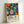 May include: Framed poster print featuring colorful abstract botanical artwork with vibrant flowers, leaves, and organic shapes in orange, blue, pink, yellow, and green against black dotted background. Text shows "Yayoi Kusama" and Japanese characters below the artwork. Light wooden frame leans against white wall on light surface.