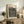 May include: Framed art print with orange "YAYOI KUSAMA" text and black polka dot pattern on cream background, displayed on rustic wooden sideboard alongside fern plant, ceramic vase, and books in neutral-toned living room setting with beige armchair.