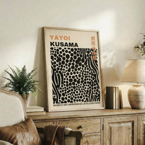 May include: Framed art print with orange "YAYOI KUSAMA" text and black polka dot pattern on cream background, displayed on rustic wooden sideboard alongside fern plant, ceramic vase, and books in neutral-toned living room setting with beige armchair.
