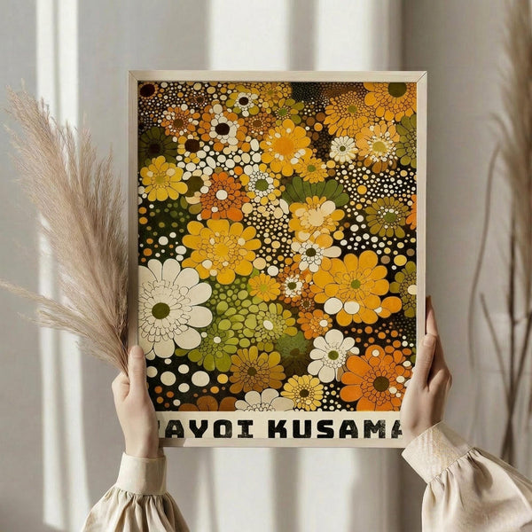 May include: Person holding framed art print featuring vibrant floral pattern with dots and circles in yellow, orange, green, white and brown tones against dark background. Hands visible wearing cream colored ruffle-sleeve top, holding dried pampas grass stems. Soft natural lighting with white curtains in background. Print has light wooden frame and white border with black text at bottom.