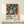 May include: Framed colorful botanical art print with abstract floral patterns in blues, oranges, pinks and yellows on black background, displayed on light wood console table against cream wall. Small white wrapped gift box with evergreen sprig sits in front of frame. Artwork features polka dots, leaves, and organic shapes in vibrant contemporary style within thin natural wood frame.