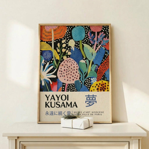May include: Framed colorful botanical art print with abstract floral patterns in blues, oranges, pinks and yellows on black background, displayed on light wood console table against cream wall. Small white wrapped gift box with evergreen sprig sits in front of frame. Artwork features polka dots, leaves, and organic shapes in vibrant contemporary style within thin natural wood frame.