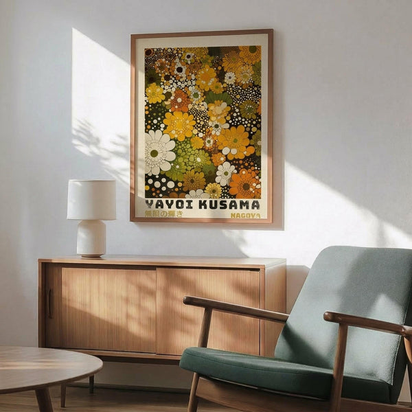 May include: Colorful floral poster with vibrant yellow, orange, green, and white dotted flower patterns in wooden frame, displayed above natural wood sideboard with white ceramic lamp. Mid-century modern sage green armchair with wooden arms sits nearby in bright, sunlit room with white walls and warm shadows.