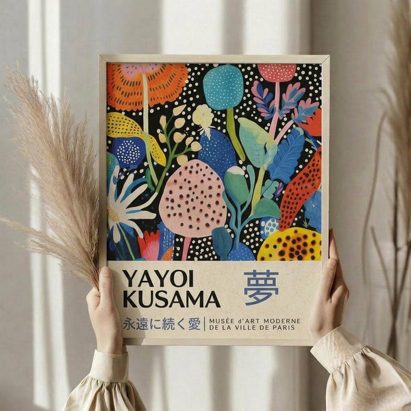 May include: Person holding framed colorful abstract botanical art print featuring stylized flowers, leaves, and circular shapes in vibrant blues, oranges, pinks, and yellows with polka dot patterns on black background. Text shows "YAYOI KUSAMA" and Japanese characters. Light wood frame, neutral curtain backdrop, dried wheat stems visible on left side.