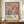 May include: Framed art poster featuring stylized floral pattern in soft pastels - pink, sage green, coral, and cream flowers with radiating petals against dark navy background. Text reads "YAYOI KUSAMA" with Japanese characters and "永遠に続く愛 MUSÉE d'ART MODERNE DE LA VILLE DE PARIS" below. Wooden frame sits on credenza next to white ceramic vase and stacked books in neutral-toned room with crown molding.