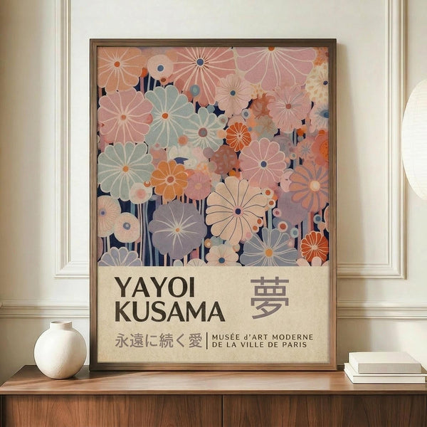 May include: Framed art poster featuring stylized floral pattern in soft pastels - pink, sage green, coral, and cream flowers with radiating petals against dark navy background. Text reads "YAYOI KUSAMA" with Japanese characters and "永遠に続く愛 MUSÉE d'ART MODERNE DE LA VILLE DE PARIS" below. Wooden frame sits on credenza next to white ceramic vase and stacked books in neutral-toned room with crown molding.