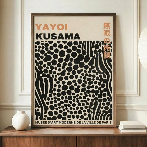 May include: Framed art poster featuring black polka dots and wavy stripes on cream background, displayed on wooden sideboard next to small white ceramic vase and stacked books. Poster shows "YAYOI KUSAMA" in orange and black text with Japanese characters, "MUSEE D'ART MODERNE DE LA VILLE DE PARIS" at bottom. Thin wooden frame against white paneled wall in bright interior setting.