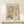 May include: Framed poster print with abstract colorful design featuring flowing organic shapes in turquoise, pink, coral, and yellow with scattered black and colored dots throughout. Text reads "YAYOI KUSAMA" and "MUSEE D'ART MODERNE DE LA VILLE DE PARIS" with Japanese characters. Light wood frame displayed on white wooden dresser beside small white gift box with greenery sprig against cream wall.