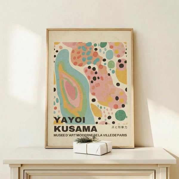 May include: Framed poster print with abstract colorful design featuring flowing organic shapes in turquoise, pink, coral, and yellow with scattered black and colored dots throughout. Text reads "YAYOI KUSAMA" and "MUSEE D'ART MODERNE DE LA VILLE DE PARIS" with Japanese characters. Light wood frame displayed on white wooden dresser beside small white gift box with greenery sprig against cream wall.