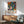 May include: Colorful Yayoi Kusama-inspired poster in thin wooden frame hanging on white wall above mid-century modern wooden credenza. Artwork features vibrant botanical motifs with polka dots in blues, oranges, pinks, and greens on black background. Room scene shows mint green upholstered chair with wooden arms, white ceramic table lamp, and natural sunlight creating shadows on wall.