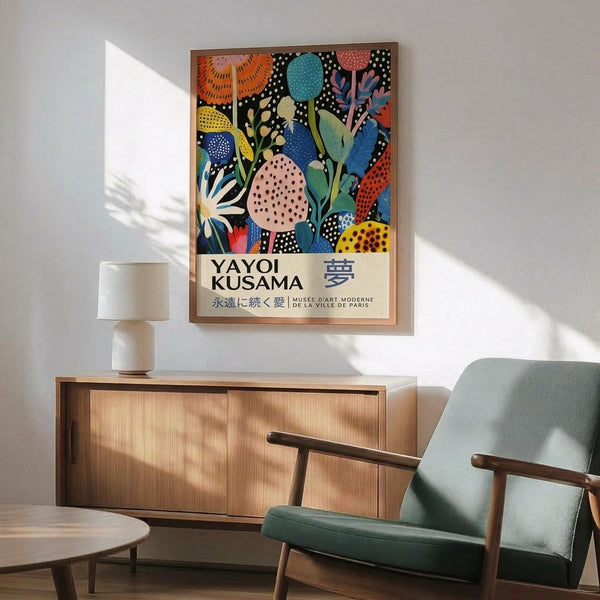 May include: Colorful Yayoi Kusama-inspired poster in thin wooden frame hanging on white wall above mid-century modern wooden credenza. Artwork features vibrant botanical motifs with polka dots in blues, oranges, pinks, and greens on black background. Room scene shows mint green upholstered chair with wooden arms, white ceramic table lamp, and natural sunlight creating shadows on wall.