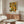 May include: Framed poster featuring colorful floral pattern with yellow, orange, green and white blooms and polka dots on dark background, displayed above wooden sideboard in modern living room with white curved chair, glass coffee table, and table lamp