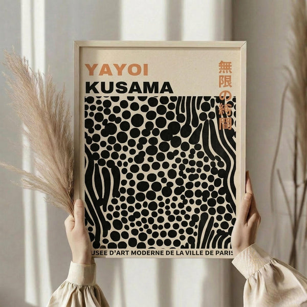 May include: Person in beige clothing holding framed poster with black polka dots and wavy lines on cream background, featuring "YAYOI KUSAMA" text in orange and black lettering with Japanese characters. Light wood frame, neutral room setting with pampas grass visible to left, soft natural lighting through white curtains.