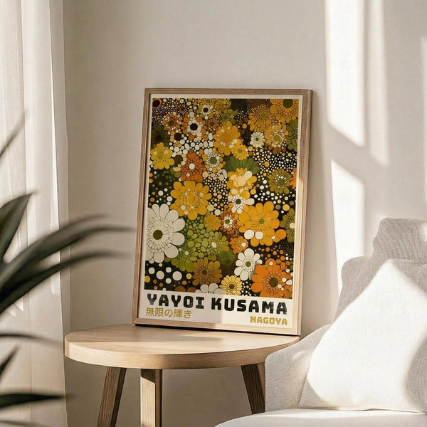 May include: Framed poster with colorful floral pattern in yellow, orange, green, and white tones displayed on light wood round side table next to beige upholstered chair. Sunlight streams across white wall creating soft shadows. Green plant leaves visible in corner. Artwork features dense arrangement of stylized flowers and polka dots in retro 1970s aesthetic with Japanese text at bottom.