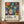 May include: Framed art poster featuring vibrant botanical illustration with colorful abstract flowers, leaves, and organic shapes in blues, oranges, pinks, and yellows against black polka-dotted background. Text reads "YAYOI KUSAMA" with Japanese characters and "MUSÉE D'ART MODERNE DE LA VILLE DE PARIS" below. Displayed on wooden sideboard next to white ceramic vase and stacked books in room with cream walls and white trim molding.