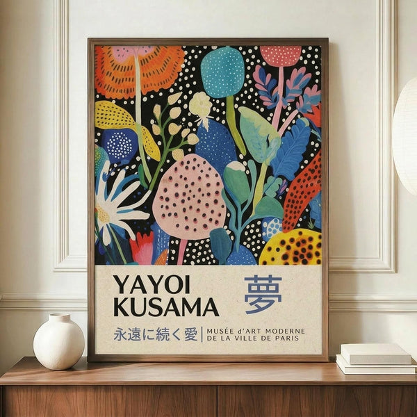 May include: Framed art poster featuring vibrant botanical illustration with colorful abstract flowers, leaves, and organic shapes in blues, oranges, pinks, and yellows against black polka-dotted background. Text reads "YAYOI KUSAMA" with Japanese characters and "MUSÉE D'ART MODERNE DE LA VILLE DE PARIS" below. Displayed on wooden sideboard next to white ceramic vase and stacked books in room with cream walls and white trim molding.