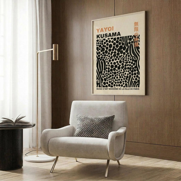 May include: Framed Yayoi Kusama exhibition poster with black polka dot pattern and flowing lines on cream background, displayed above beige upholstered armchair with geometric patterned pillow. Modern reading corner features brass floor lamp, dark side table with open book, wood paneled wall, and natural lighting from nearby window with curtains.