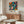 May include: Colorful abstract floral poster featuring vibrant botanical shapes in blues, oranges, pinks and yellows with polka dot patterns, displayed in thin frame on wooden sideboard beside white sculptural lamp and round mirror in modern living room with cream chair and glass coffee table.