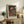 May include: Framed poster featuring colorful mushrooms with polka-dotted caps in vibrant reds, oranges, and blues against a dark starry background, displayed on a rustic wooden sideboard alongside books, a ceramic vase, and a fern plant in a cozy living room setting with neutral walls and brown leather furniture.