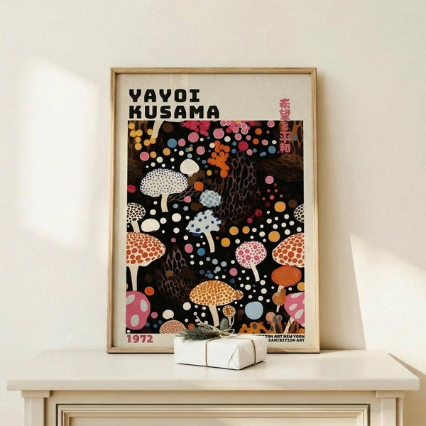 May include: Framed poster with black background featuring colorful polka-dotted mushrooms in various sizes and patterns. White, pink, orange, and blue spotted fungi scattered across dark surface with floating circles. Text reads "YAYOI KUSAMA" and "1972" with Japanese characters. Natural wood frame displayed on light wooden surface next to small white wrapped gift with dried plant sprig.