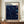 May include: Framed art poster displayed on wooden sideboard showing swirling dark blue cosmic pattern with colorful dots resembling planets and stars. White matted frame sits against cream wainscoted wall beside white ceramic vase and stacked books. Text reads "YAYOI KUSAMA" at top with Japanese characters and "1991" at bottom. Rich navy background features flowing organic lines dotted with blue, yellow and white spheres creating celestial movement.