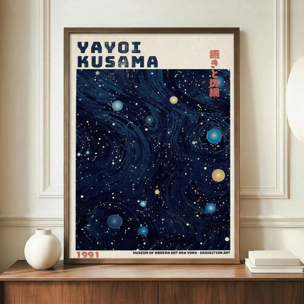 May include: Framed art poster displayed on wooden sideboard showing swirling dark blue cosmic pattern with colorful dots resembling planets and stars. White matted frame sits against cream wainscoted wall beside white ceramic vase and stacked books. Text reads "YAYOI KUSAMA" at top with Japanese characters and "1991" at bottom. Rich navy background features flowing organic lines dotted with blue, yellow and white spheres creating celestial movement.