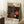 May include: Framed exhibition poster featuring Yayoi Kusama's distinctive polka dot artwork in black background with red, orange, and cream colored circles and flowing dotted patterns. Text shows "YAYOI KUSAMA," "TOKYO 1998," and "22-APRIL 1986" with Japanese characters. Poster leans against white wall corner next to coffee cup on wooden block and beige fabric, displaying vintage museum exhibition design with cream matting and natural wood frame.