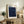 May include: Framed poster with deep blue starry night design featuring swirling patterns and colorful planets, displayed on rustic wooden sideboard alongside fern plant, ceramic vase, and books in modern living room with cream armchair and neutral walls