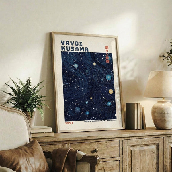 May include: Framed poster with deep blue starry night design featuring swirling patterns and colorful planets, displayed on rustic wooden sideboard alongside fern plant, ceramic vase, and books in modern living room with cream armchair and neutral walls