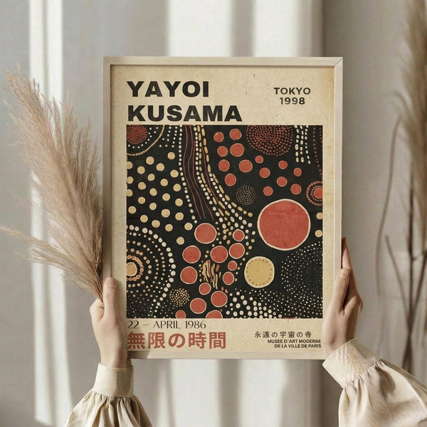May include: Person in beige clothing holding framed poster with abstract polka dot pattern in black, red, orange and cream colors, alongside dried pampas grass stems against white curtained background. Frame appears light wood with cream matting, poster features Japanese text and museum information.