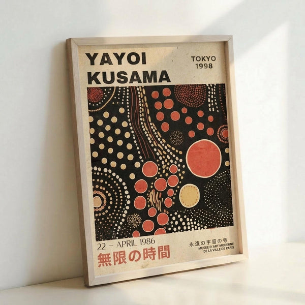 A framed exhibition poster featuring Yayoi Kusama's distinctive polka dot artwork on a black background with red, orange, and cream colored circles in various sizes. The poster displays "YAYOI KUSAMA" at the top, "TOKYO 1998" in the upper right, and Japanese text at the bottom with "22 - APRIL 1986". The artwork shows flowing, organic patterns of dots creating cosmic, infinity-net style compositions. The poster is mounted in a light wooden frame and photographed leaning aga