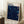 May include: Framed art print leaning against white wall corner on textured cream rug. Poster features deep navy blue swirling patterns with scattered white dots and colorful circular orbs in blues, yellow, and teal. Text reads "YAYOI KUSAMA" at top and "1991 MUSEUM OF MODERN ART NEW YORK EXHIBITION ART" at bottom. Light wood frame, coffee cup on wooden block nearby, beige throw blanket visible.