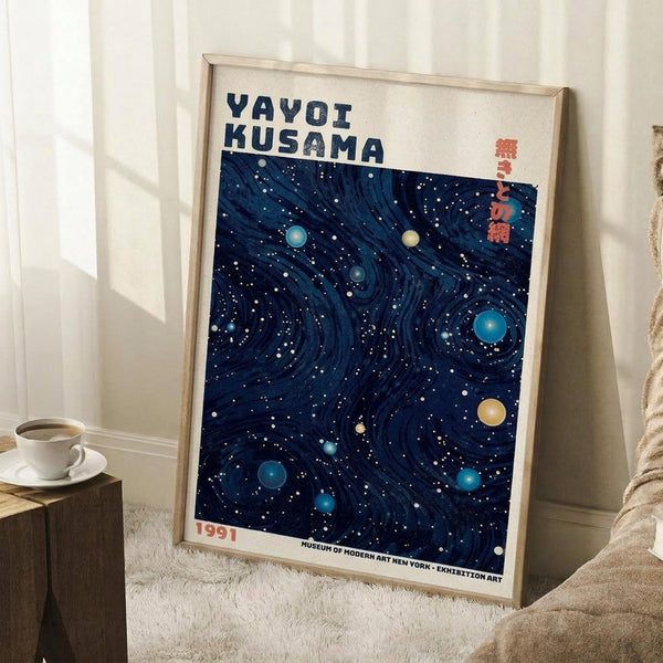 May include: Framed art print leaning against white wall corner on textured cream rug. Poster features deep navy blue swirling patterns with scattered white dots and colorful circular orbs in blues, yellow, and teal. Text reads "YAYOI KUSAMA" at top and "1991 MUSEUM OF MODERN ART NEW YORK EXHIBITION ART" at bottom. Light wood frame, coffee cup on wooden block nearby, beige throw blanket visible.